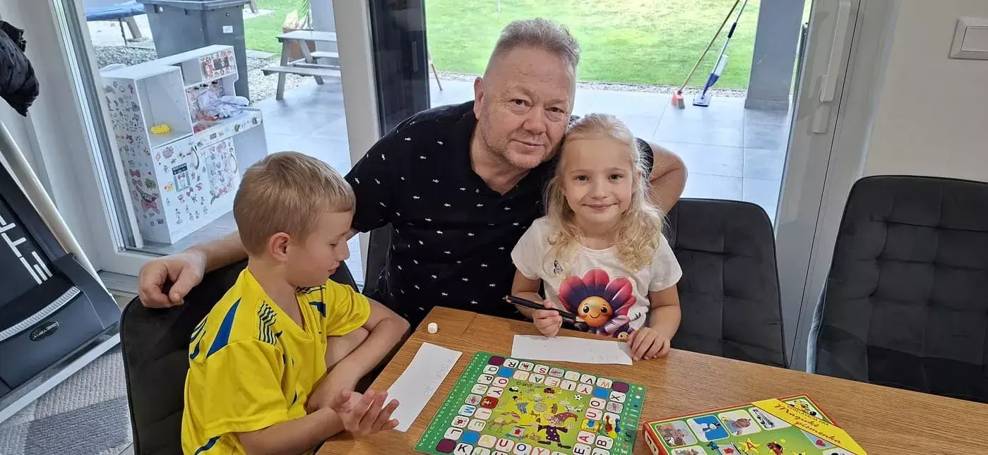 Dedko objíma dve vnúčatá sediace pri hracej doske Magických písmen s otvorenou krabicou hry Miaplay