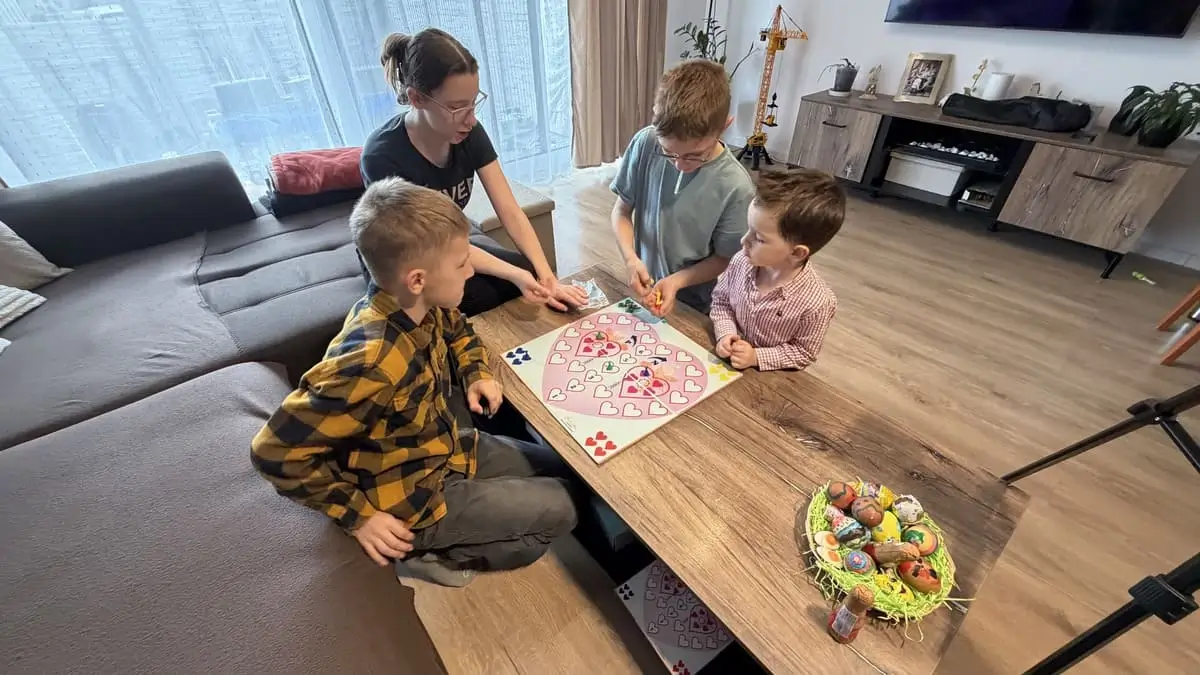 Štyrai súrodenci rôzneho veku hrajú spoločenskú hru Srdiečka pri rodinnom stole v obývačke