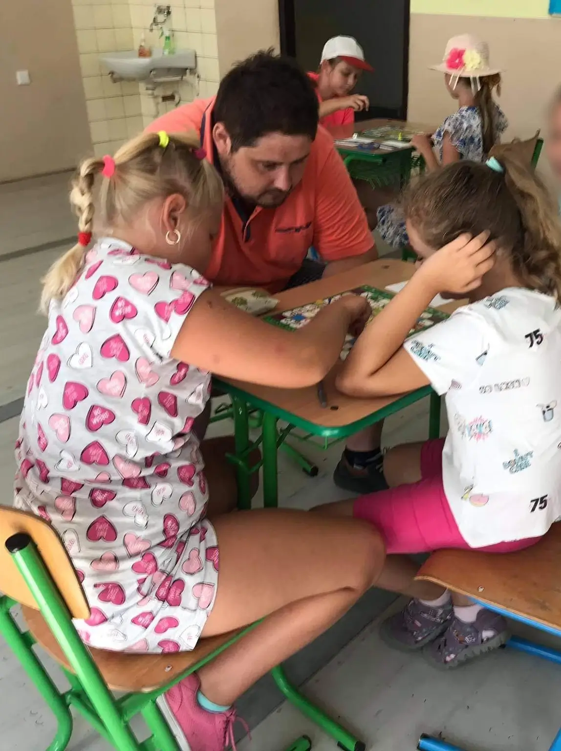 Učiteľ v oranžovom tričku pomáha dvom dievčatám pri spoločenskej hre Miaplay na letnom detskom tábore